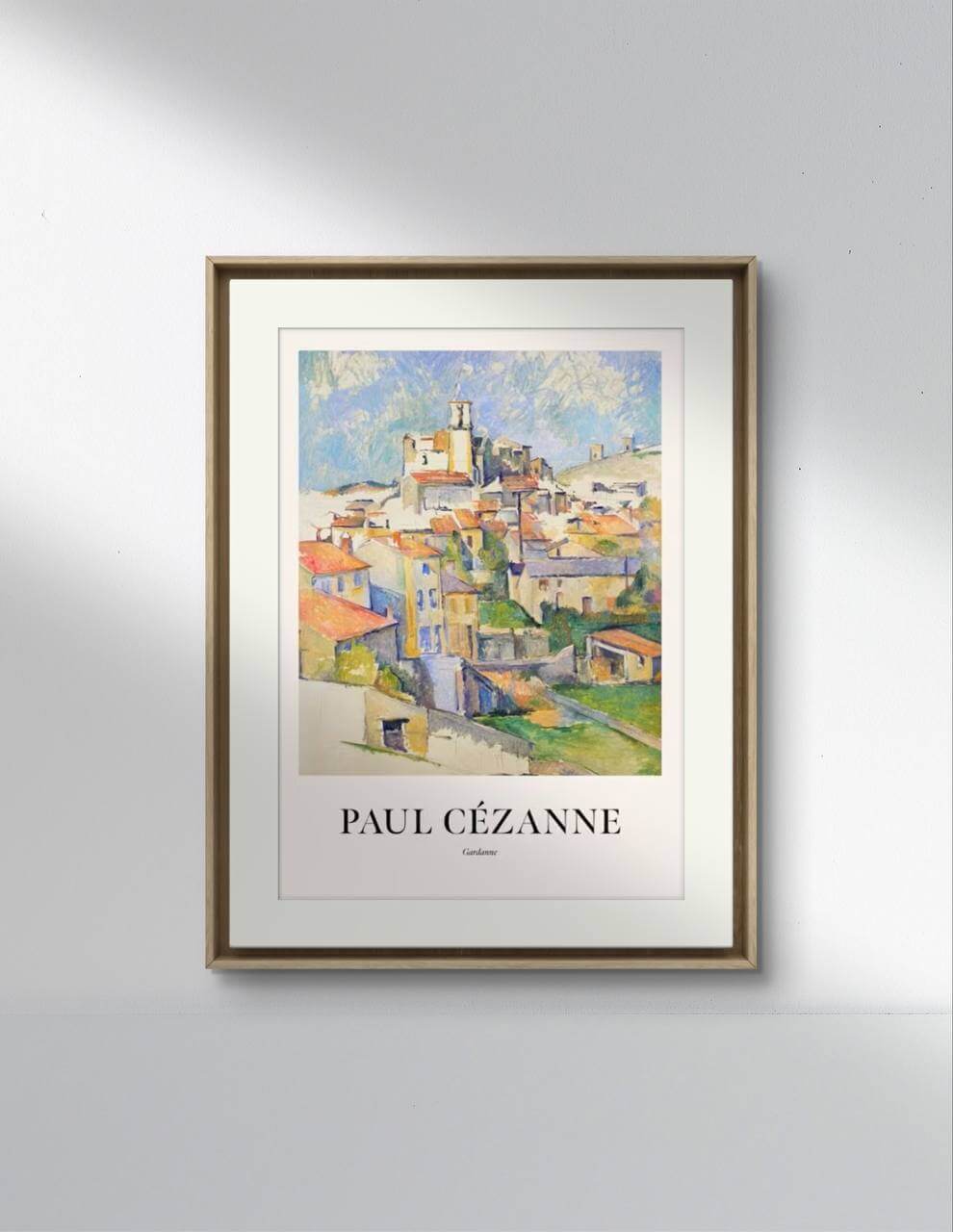 A modern living room with a light gray sofa and a beige knitted blanket. On the wall, above a low bookshelf with books, hangs the poster of the painting "Gardanne" by Paul Cézanne, framed and serving as the focal point of the decor. A small, modern table lamp is on the right of the poster.