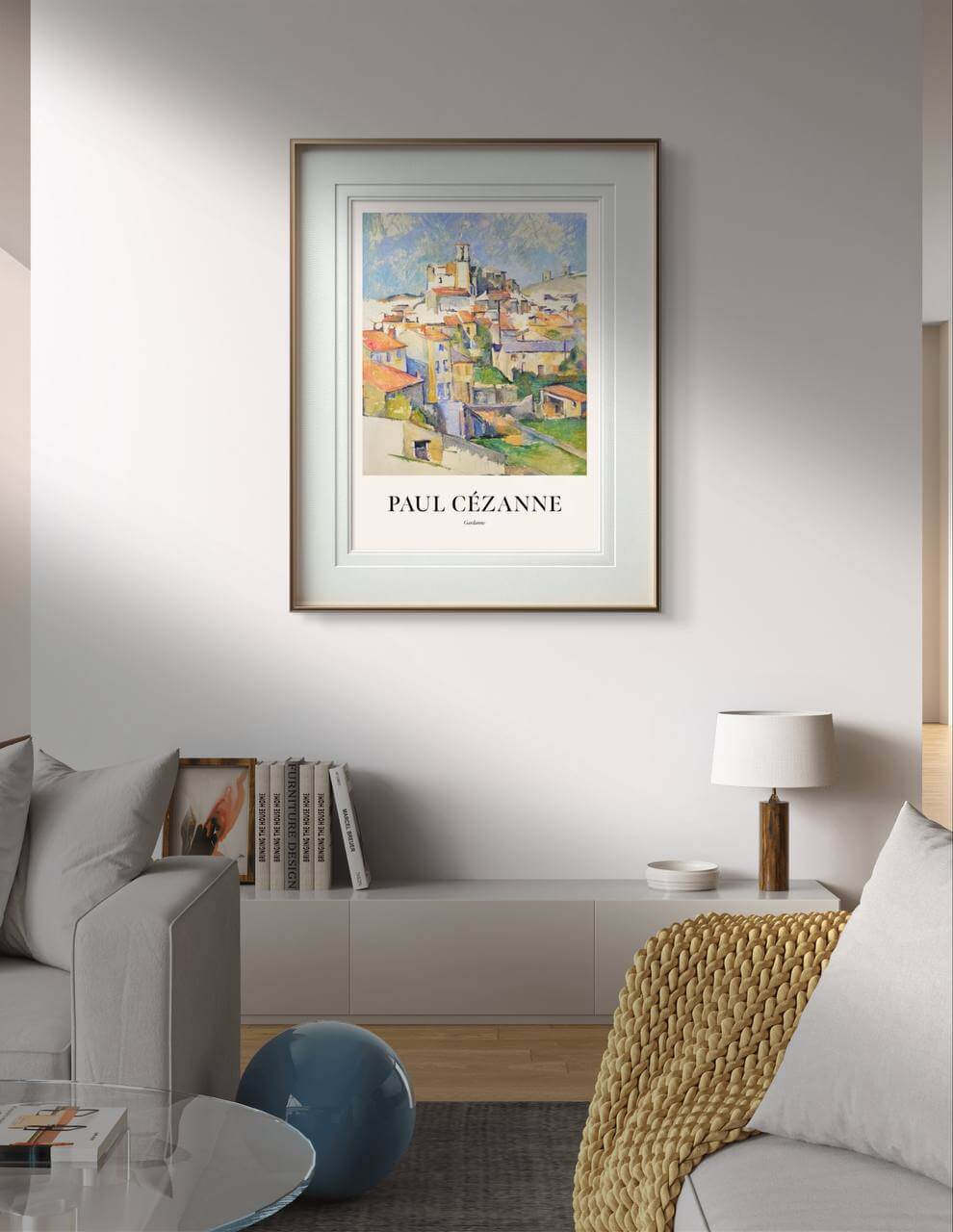 A modern living room with a light gray sofa and a beige knitted blanket. On the wall, above a low bookshelf with books, hangs the poster of the painting "Gardanne" by Paul Cézanne, framed and serving as the focal point of the decor. A small, modern table lamp is on the right of the poster. This poster print is perfect for home decor, bringing classic art into your home gallery wall.
