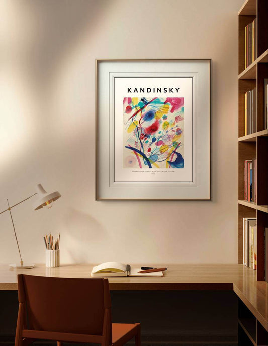 A contemporary study space with a wooden desk, a modern lamp, and a chair. The poster of "Composition in Red, Blue, Green, and Yellow" by Kandinsky is framed and hung on the wall above the desk. Bookshelves filled with books are on the right. This painting reproduction serves as excellent wall art for a home gallery wall or a stylish office decor