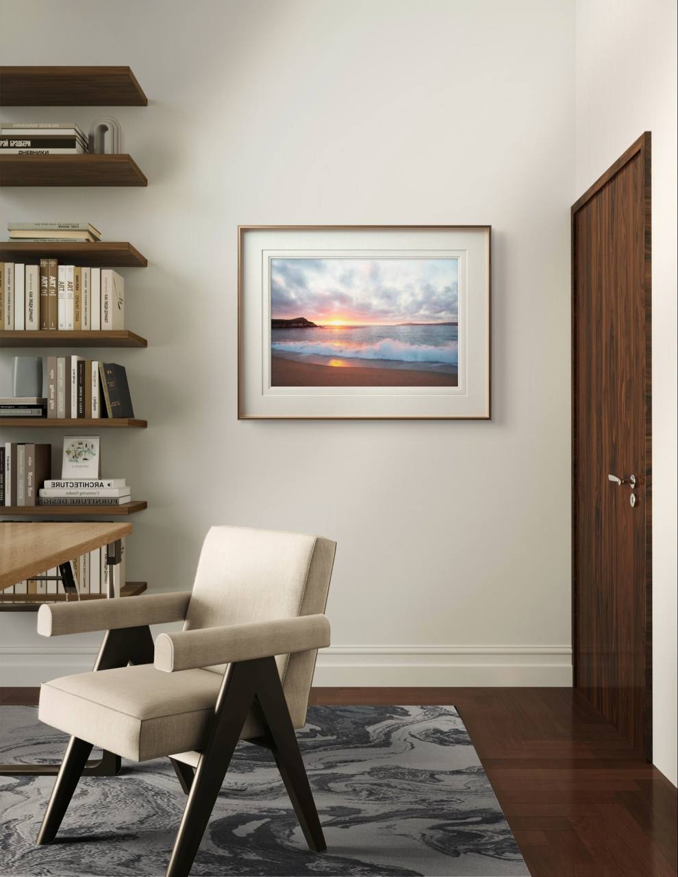 A framed beach sunset poster hangs on a light-colored wall in a modern room. A beige armchair, wooden shelves, and books complete the warm, cozy ambiance of the space.