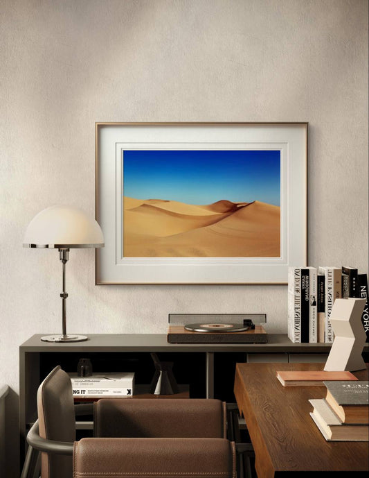A framed sand dunes poster hangs above a desk in a stylish study. The space features a turntable, books, and a lamp, offering a warm and sophisticated atmosphere.