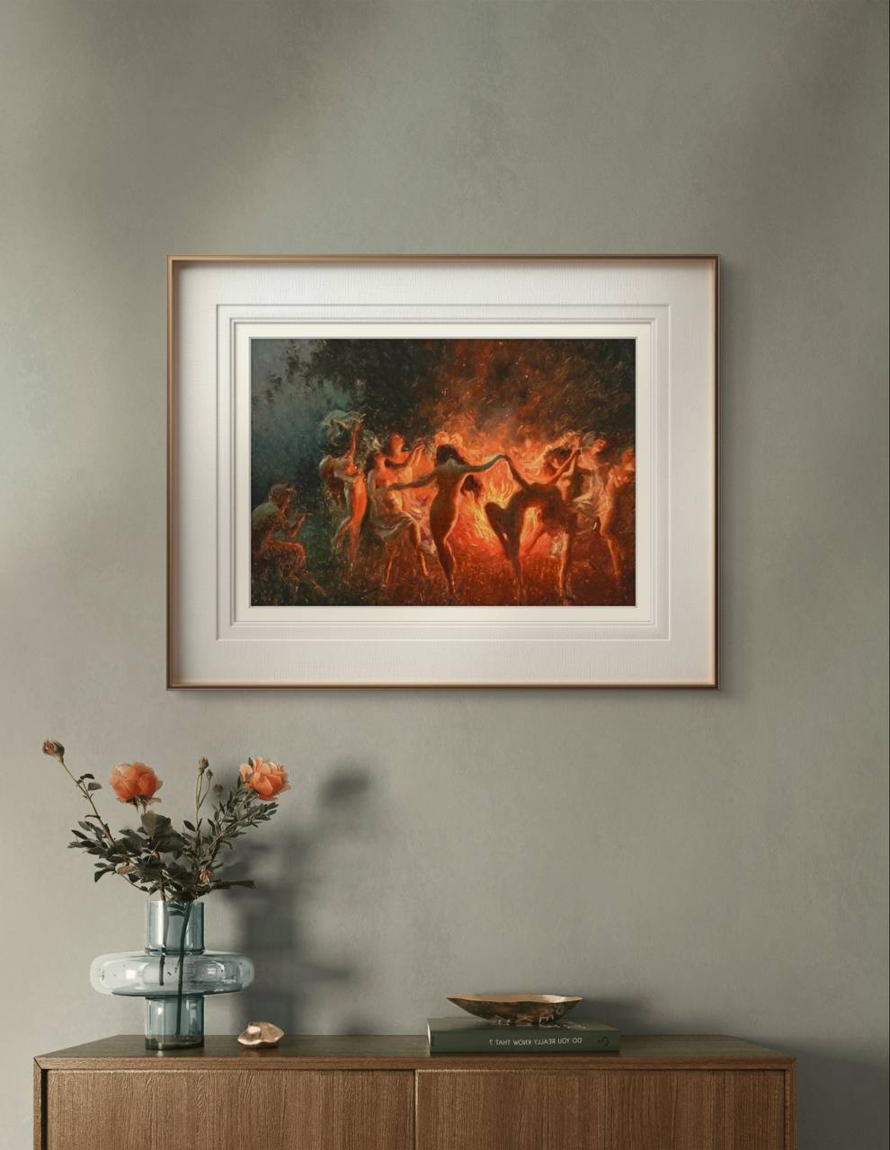 The framed painting of nymphs dancing around a bonfire is displayed in a modern room, hung above a wooden cabinet with flowers. The warm tones of the painting's flames add a dramatic touch to the neutral, minimalist decor.