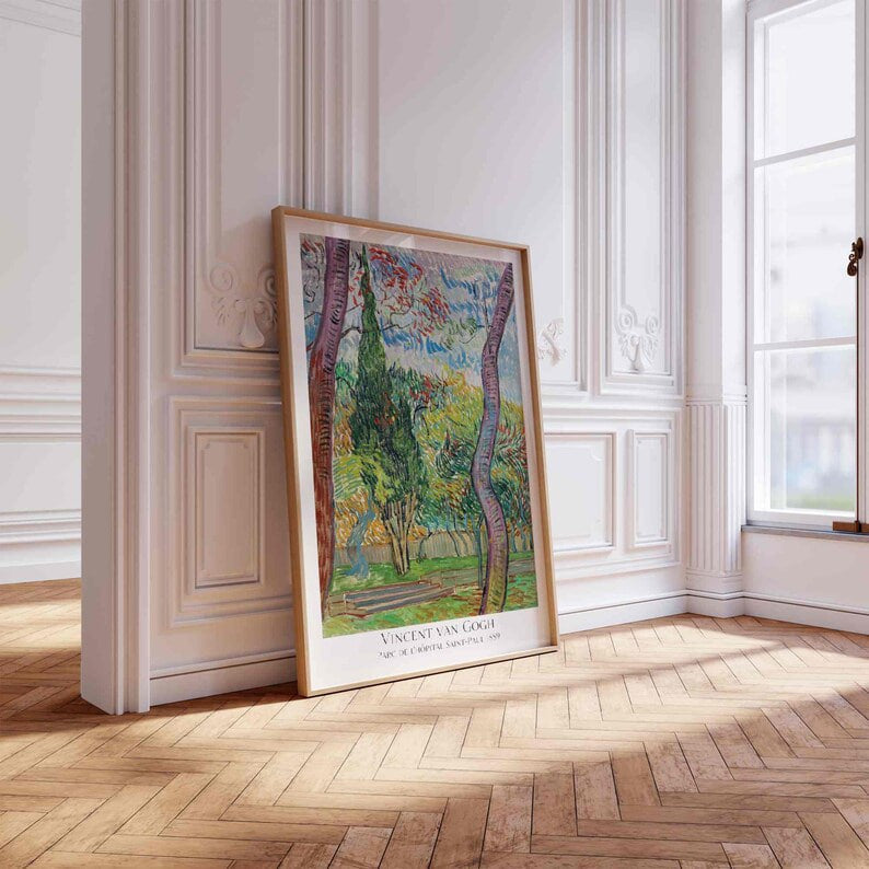 The first image shows a framed poster leaning against an ornate white wall in a bright, sunlit room. The wall features intricate moldings that add a touch of elegance. The poster is a reproduction of Vincent van Gogh's painting titled "Parc de l'Hôpital Saint-Paul, 1889." The artwork vividly captures a colorful garden scene with two tall, twisting trees framing the view. The foliage is vibrant with shades of green, red, and blue, illustrating Van Gogh's expressive brushwork. The frame itself is simple with