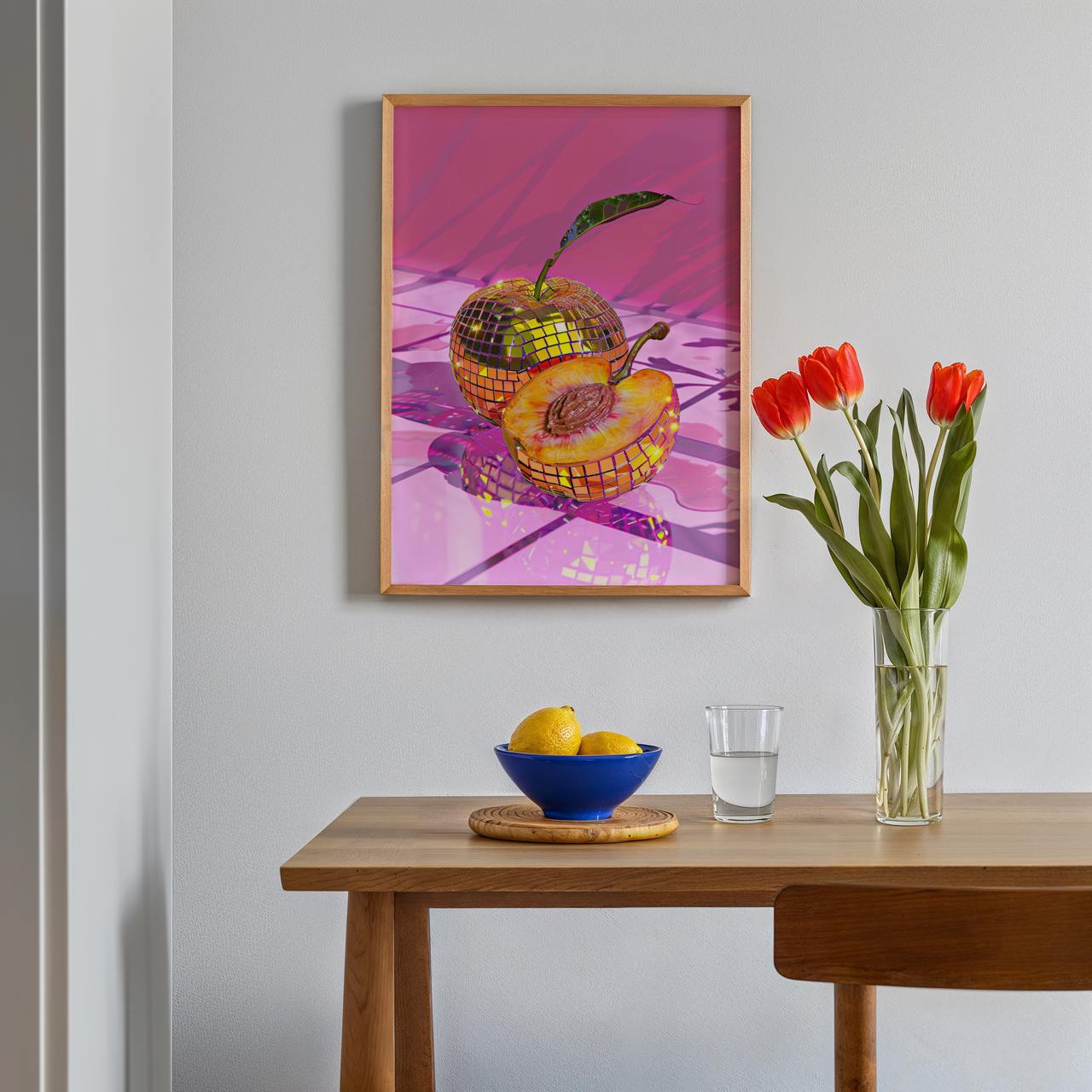 Framed artwork on a wall above a wooden table with a bowl of fruit, glass of water, and vase of flowers.