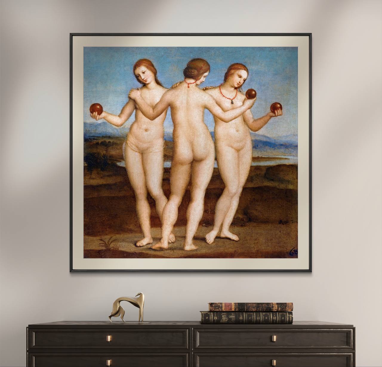 The first image displays a framed poster of Raphael's painting "Three Graces." The artwork features three nude women, standing in a circle, holding apples. Their elegant poses and serene expressions are beautifully captured against a soft, natural background. The poster is framed in a sleek black frame and hung above a dark wooden cabinet, blending classic art with contemporary decor.