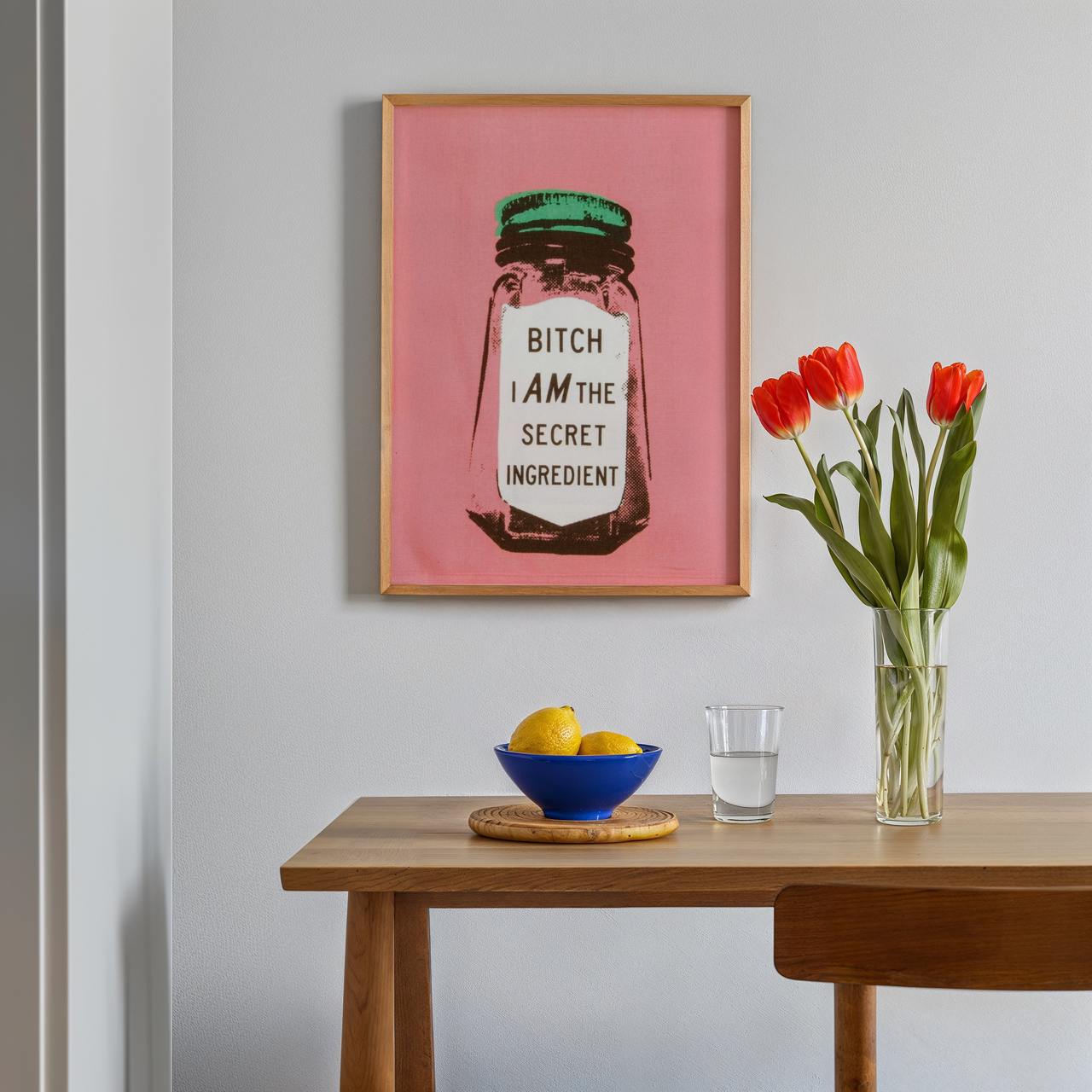 Framed artwork on a wall above a wooden table with a bowl, glass, and vase of tulips.