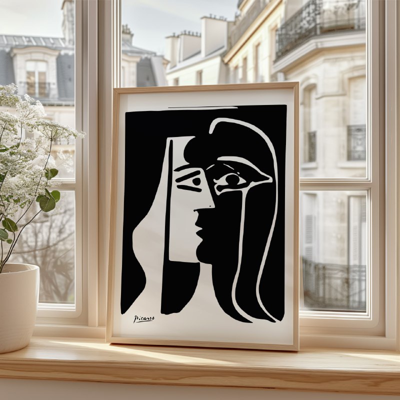 The image shows a framed black-and-white artwork in Picasso's abstract style, resting on a windowsill with Parisian buildings visible outside. The artwork features a dual-profile face in bold, minimalist lines, capturing two perspectives within one head. A white plant in a pot sits beside the frame, adding a touch of nature.