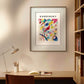 A contemporary study space with a wooden desk, a modern lamp, and a chair. The poster of "Composition in Red, Blue, Green, and Yellow" by Kandinsky is framed and hung on the wall above the desk. Bookshelves filled with books are on the right. This painting reproduction serves as excellent wall art for a home gallery wall or a stylish office decor