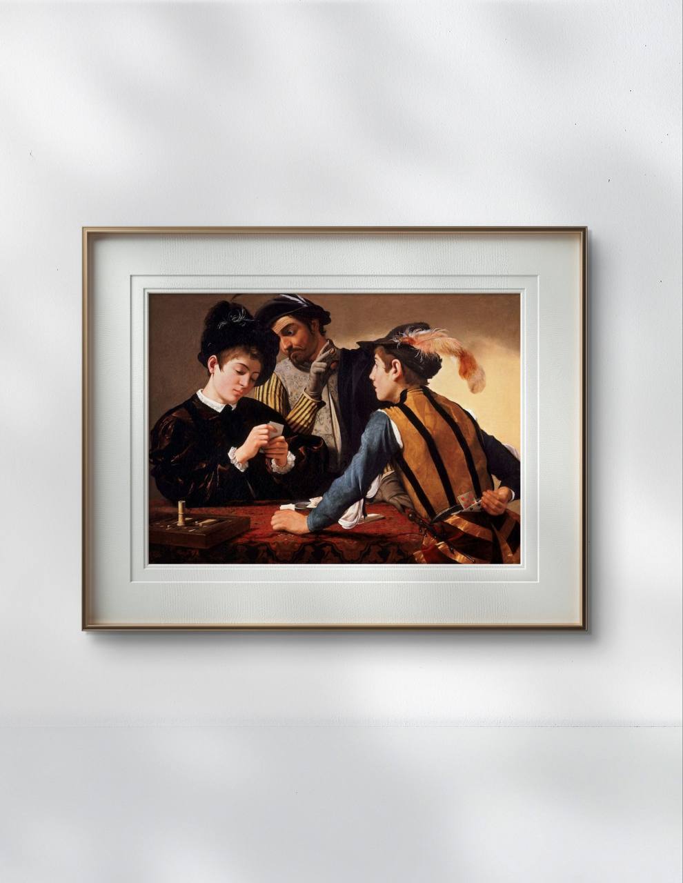 The same artwork, "The Cardsharps" by Caravaggio, is elegantly framed in white with a gold trim, hanging on a plain white wall. The scene depicted is of three men playing cards, with an air of suspense.