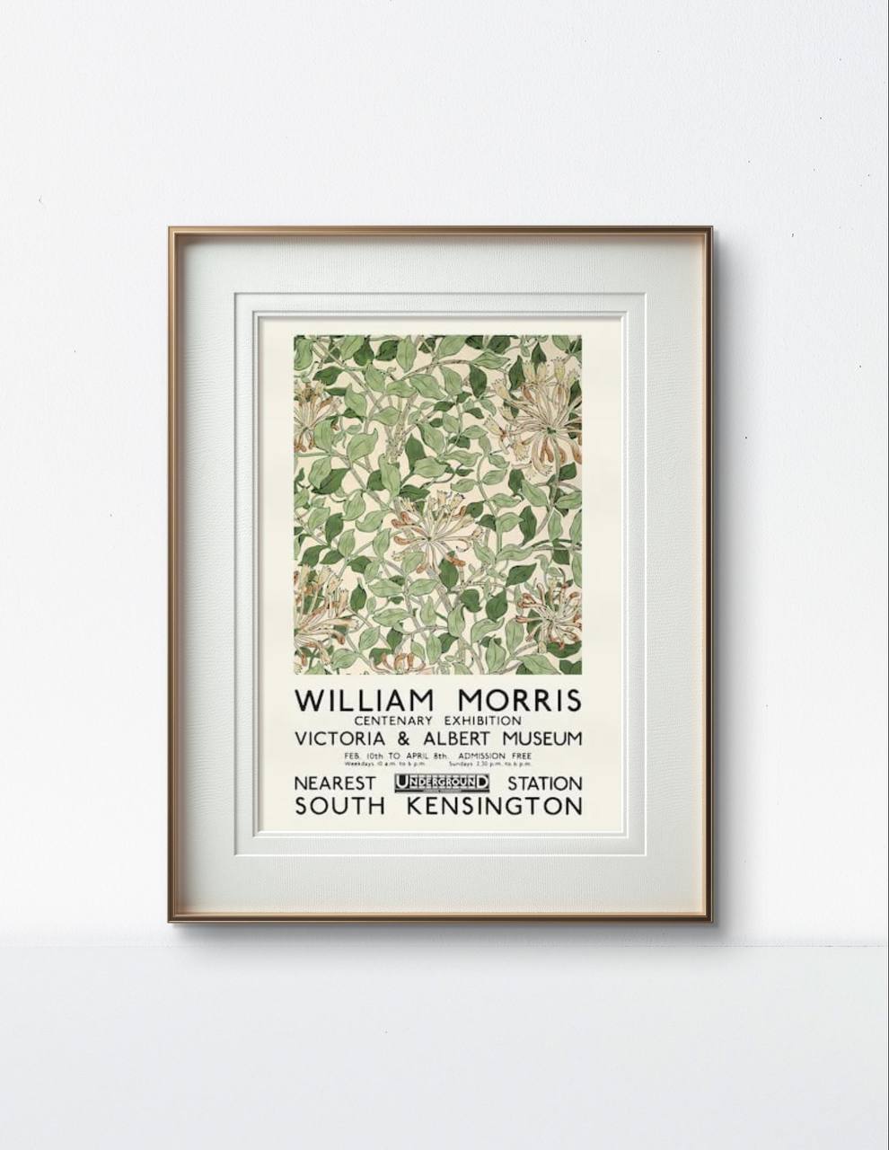 The framed William Morris exhibition poster hangs on a simple white wall. The green and orange botanical design contrasts beautifully with the neutral room, while the black text provides information about the exhibition.