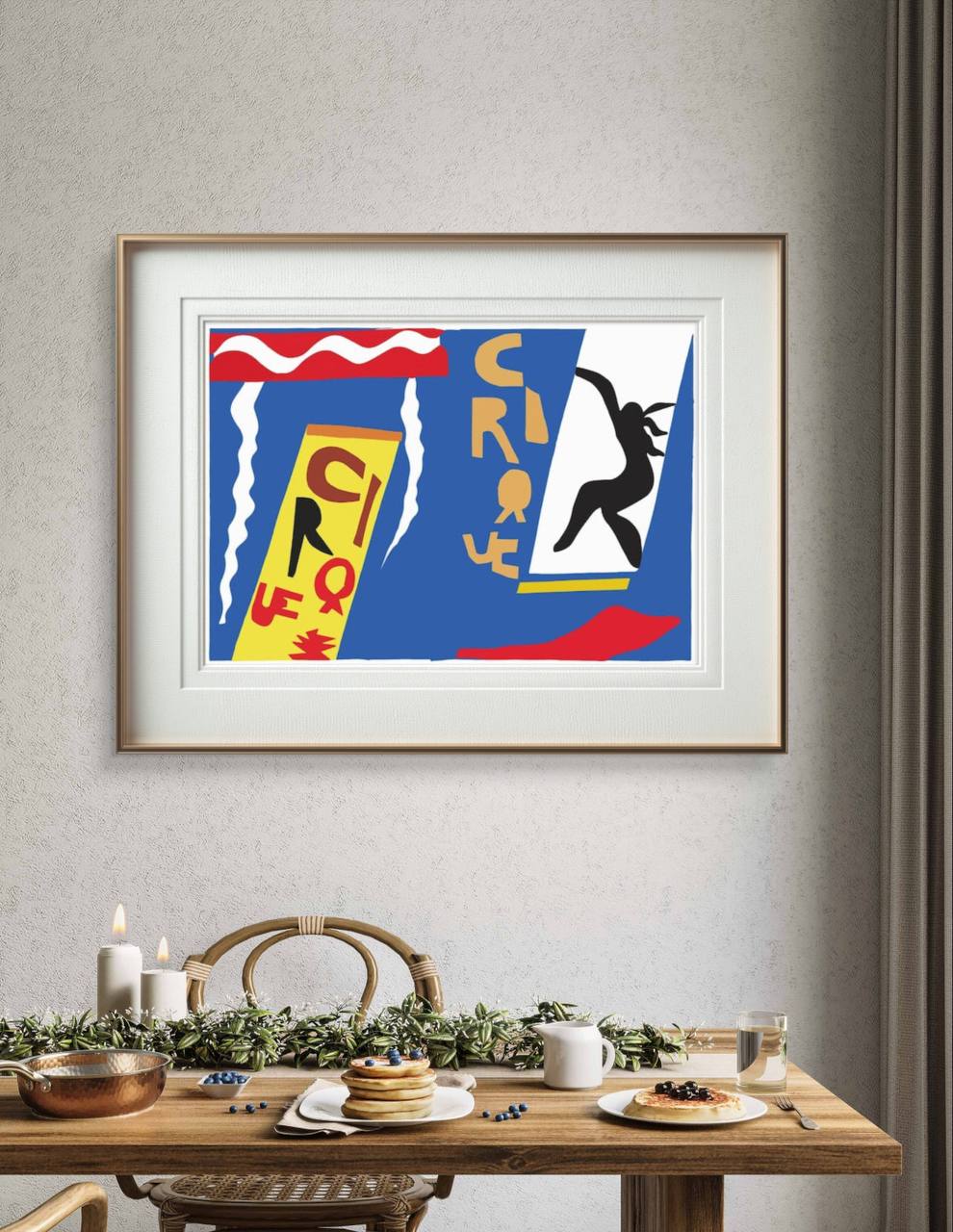 A framed abstract poster with colorful shapes and the word 'Cirque' hangs above a dining table. The room is decorated with pancakes, candles, and greenery, creating a cozy atmosphere