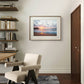 A framed beach sunset poster hangs on a light-colored wall in a modern room. A beige armchair, wooden shelves, and books complete the warm, cozy ambiance of the space.

