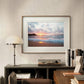 The beach sunset poster is framed and displayed above a desk in a study area. The room features books, a turntable, and a lamp, creating a sophisticated and peaceful atmosphere.