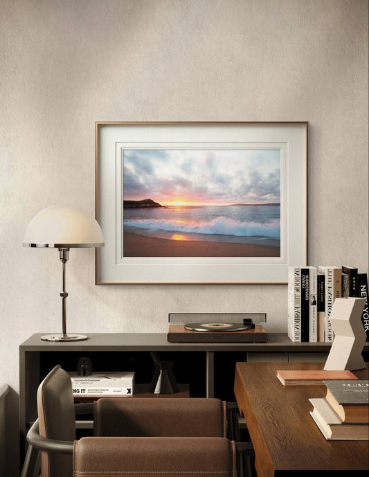 The beach sunset poster is framed and displayed above a desk in a study area. The room features books, a turntable, and a lamp, creating a sophisticated and peaceful atmosphere.