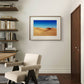 A framed sand dunes poster is displayed on a light-colored wall in a modern room. A beige armchair, wooden shelves, and books create a peaceful and contemporary environment.

