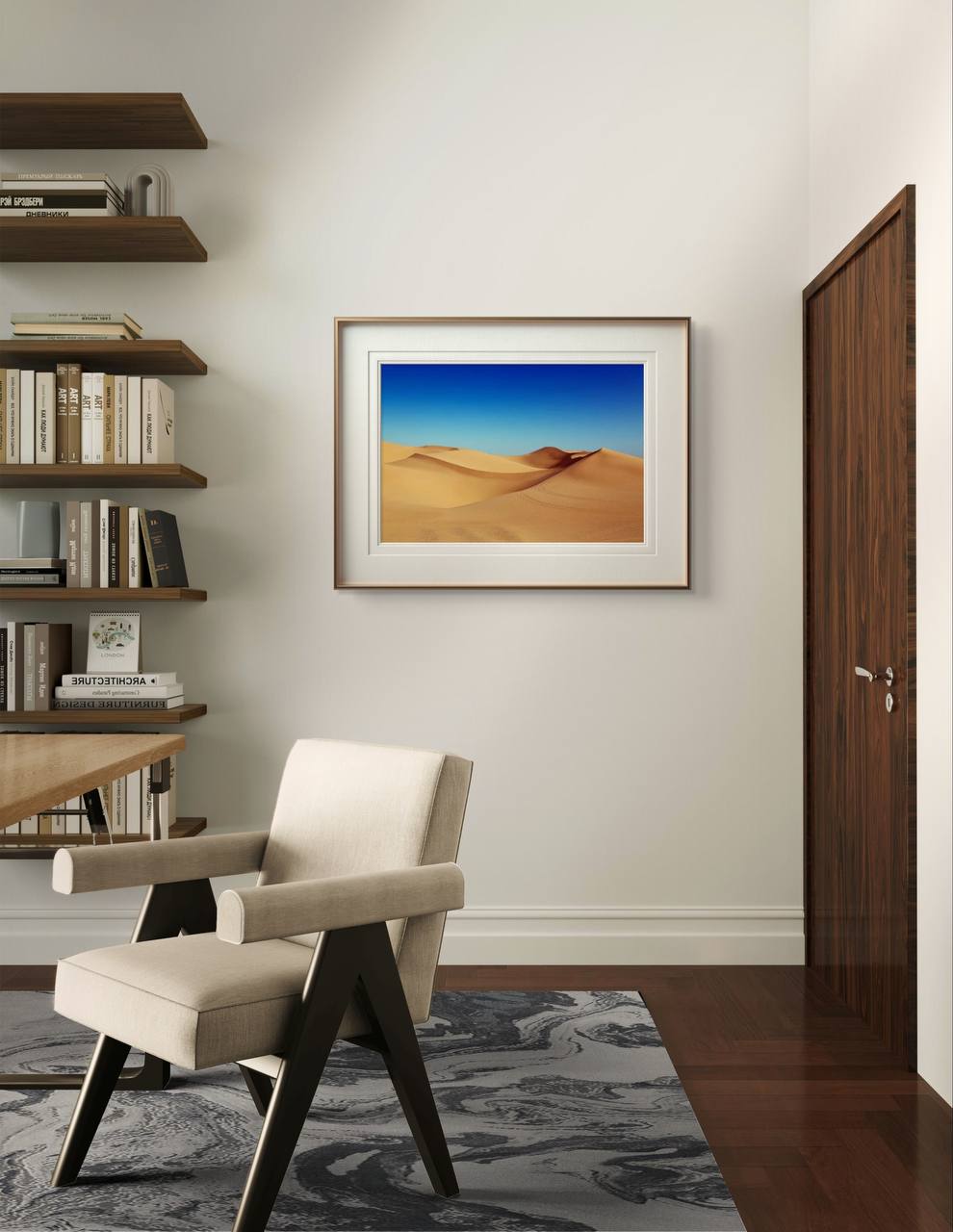 A framed sand dunes poster is displayed on a light-colored wall in a modern room. A beige armchair, wooden shelves, and books create a peaceful and contemporary environment.
