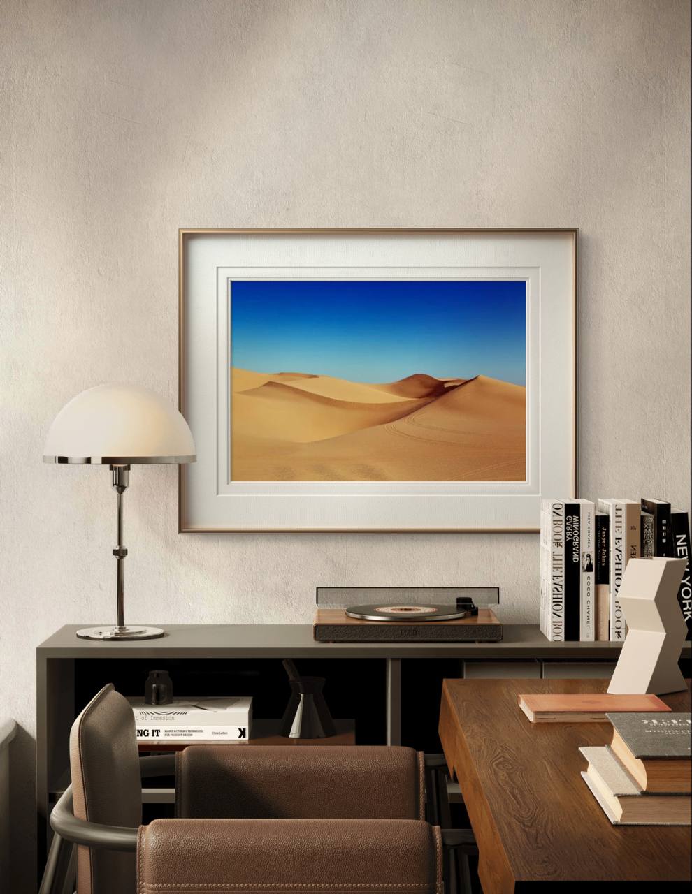 A framed sand dunes poster hangs above a desk in a stylish study. The space features a turntable, books, and a lamp, offering a warm and sophisticated atmosphere.