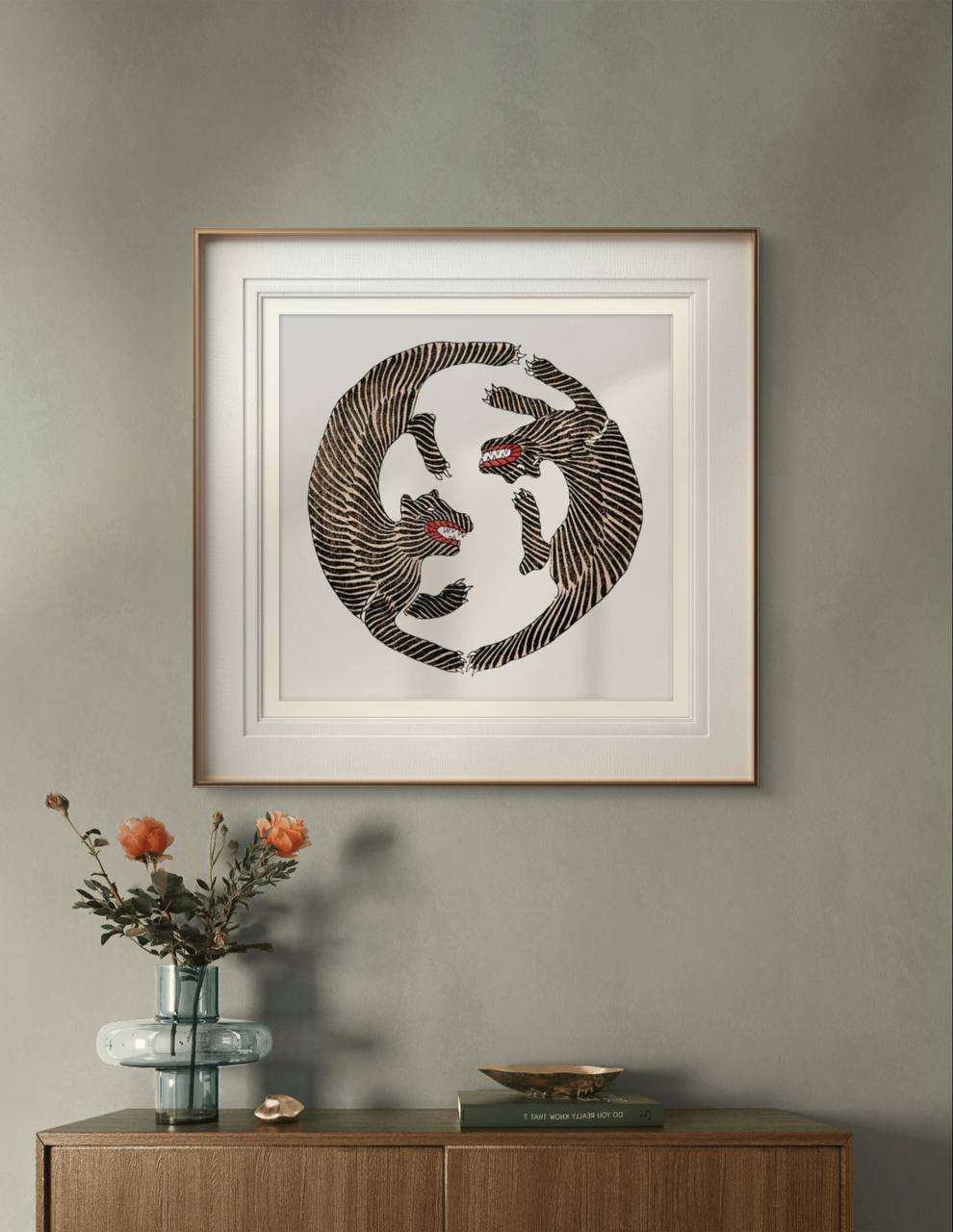 A framed illustration of two black-striped tigers, positioned in a circular pattern with red mouths open. The minimalist, elegant design is framed in gold and white, hanging above a wooden cabinet with a glass vase and orange flowers.