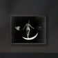 The framed artwork of the woman on the crescent moon is displayed against a dark wall. The stark contrast between the black background and the illuminated moon and figure creates a striking, mystical ambiance in the room.