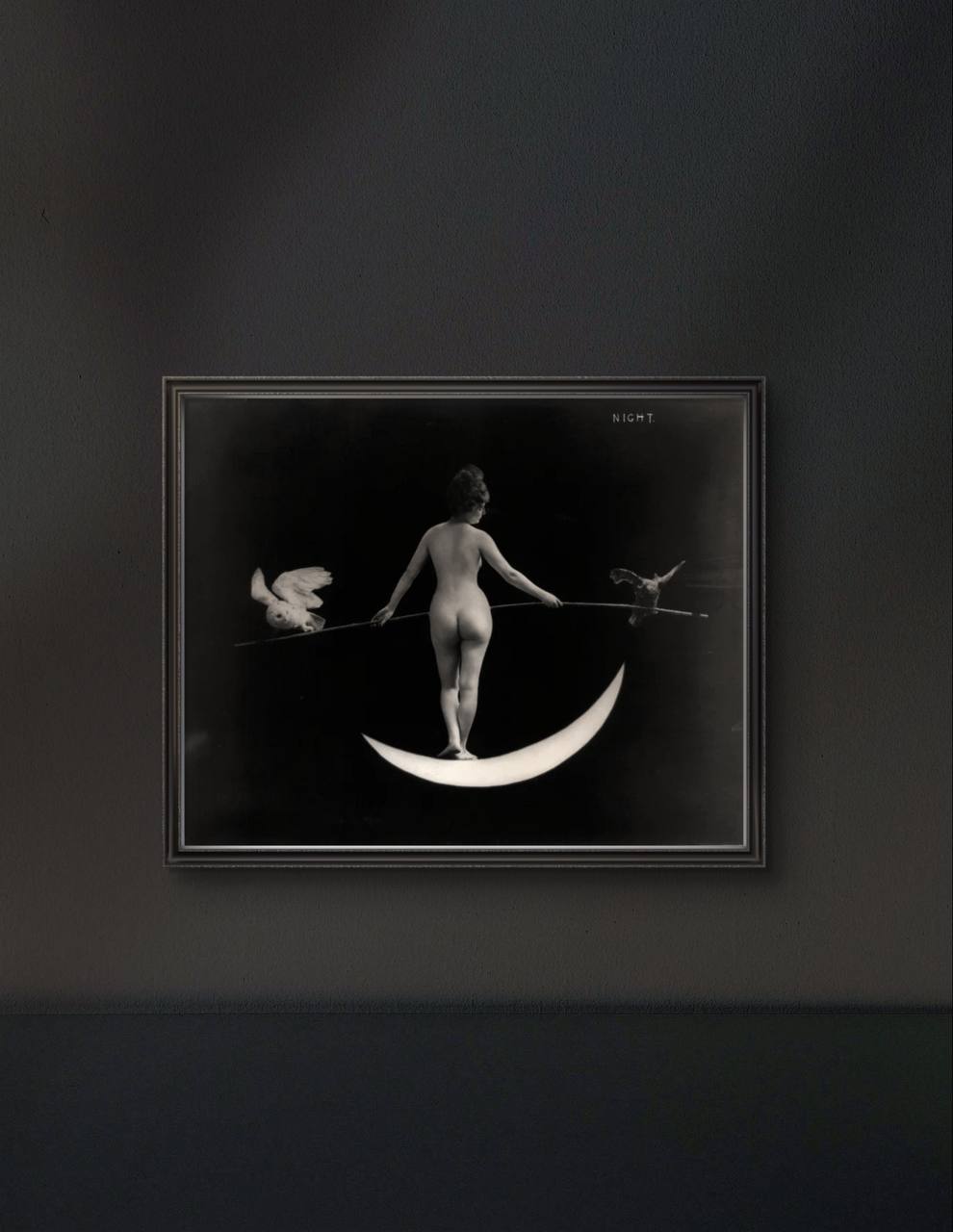 The framed artwork of the woman on the crescent moon is displayed against a dark wall. The stark contrast between the black background and the illuminated moon and figure creates a striking, mystical ambiance in the room.