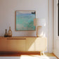 A framed painting of Lake Attersee in a modern room, hanging above a light wooden sideboard with simple decor. The vibrant turquoise water contrasts with the muted tones of the room, creating a relaxing and natural atmosphere.