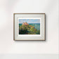 The same seaside cottage scene framed in a simple wooden frame with white matting. The vibrant greenery and calm sea contrast beautifully against the light-colored wall.