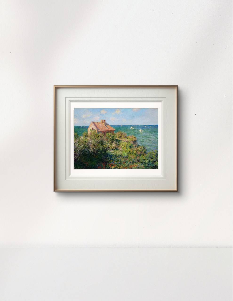 The same seaside cottage scene framed in a simple wooden frame with white matting. The vibrant greenery and calm sea contrast beautifully against the light-colored wall.