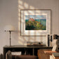 The framed artwork is displayed in a modern room, above a desk with books and a lamp. Sunlight filters through, casting soft shadows, and highlighting the serene coastal painting.