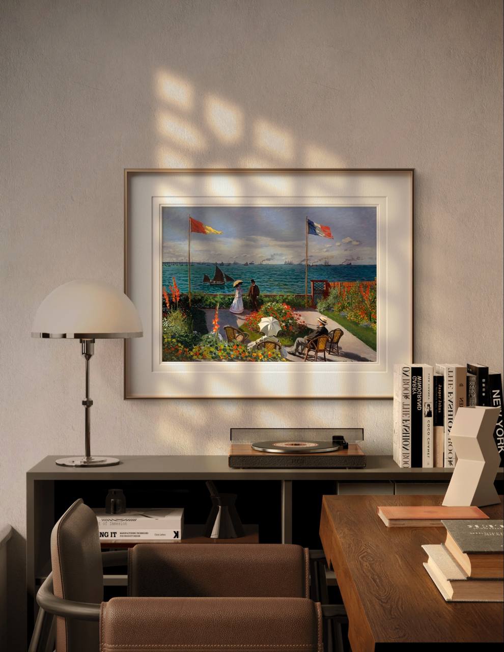 The framed painting of a coastal garden is displayed in a modern room with a desk, books, and a lamp. Natural light filters in, highlighting the serene seaside scene with flowers and boats.