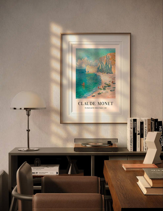 The framed beach painting is displayed above a desk, surrounded by books and a lamp. Soft sunlight fills the room, adding warmth to the tranquil scene of cliffs, sea, and boats.