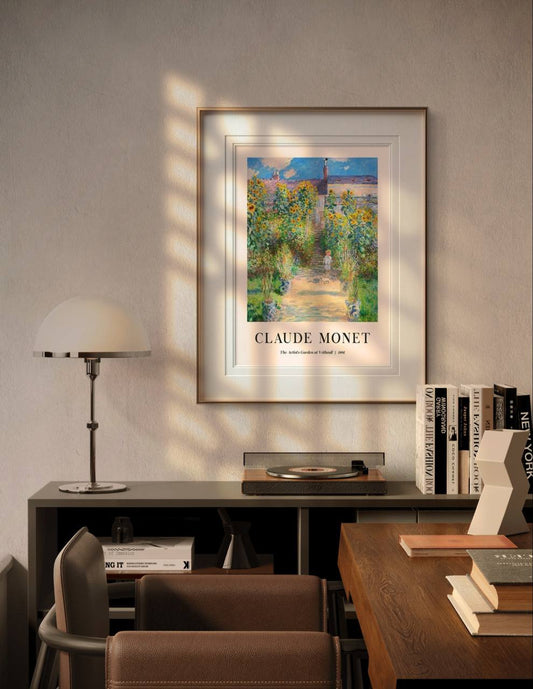 The framed painting hangs above a desk in a modern room, surrounded by books and a lamp. Soft sunlight filters in, highlighting the lush sunflowers and peaceful path in the garden.