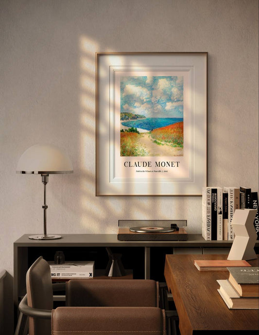 The framed painting is displayed in a modern room, above a desk and next to a lamp and books. Soft sunlight filters in, highlighting the vivid blues and warm golden tones of the landscape.