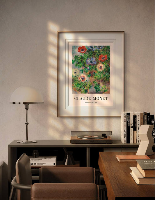 A Claude Monet poster featuring the "Anémones en Pot" painting, framed and hung above a modern desk with books and a lamp. The room is softly lit, highlighting the artwork.