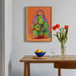 Framed artwork of a bag of apples on an orange background, displayed above a wooden table with a bowl, glass, and vase.