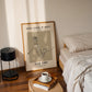 Framed artwork on a wall above a bed with a lamp and coffee cup on a side table.