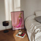 Framed artwork of a pink and gold pepper on a bedside table with a cup and book.
