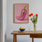 Pink embroidered artwork of a chili pepper on a wall above a wooden table with a bowl, glass, and vase.