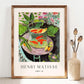 A still life painting of a glass bowl filled with bright orange goldfish, placed on a pink table, surrounded by green leaves and floral patterns. The background features lush vegetation and a decorative interior, with bold and expressive brushstrokes emphasizing color and contrast.