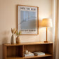 Framed poster on a wall above a wooden shelf with decor items.
