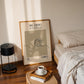 Framed artwork on a wall next to a bed with a lamp and coffee cup on a side table.