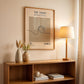 Wooden shelf with decorative items, framed artwork, and a lamp in a room setting.