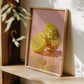 Framed artwork with a disco ball and lemon on a pink background, placed on a wooden shelf.