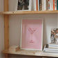 Wooden shelf with books and a framed picture of a cocktail on a pink background.
