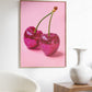 Framed artwork of pink glittery cherries on a pink background in a room with a chair and vase.