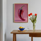 Framed artwork of a red pepper on a wall above a wooden table with a bowl, glass, and vase.