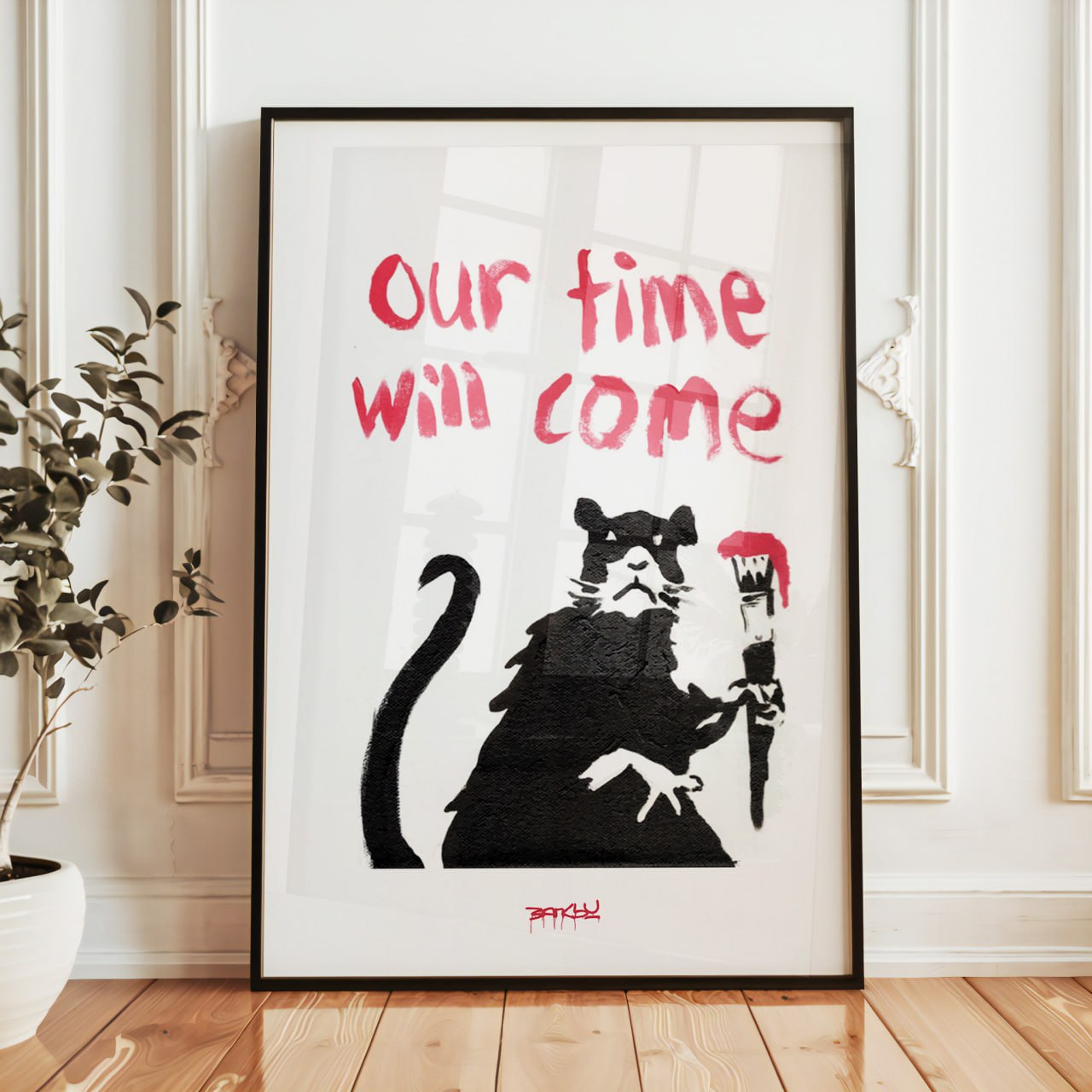 In the image, we see a framed artwork placed on the windowsill of a bright room. The artwork features a simple yet impactful design: a black rat holding a large paintbrush in its right paw, which is dripping red paint from the tip.