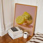 Framed artwork of a lemon and disco ball on a wall next to books and a coffee cup on a wooden floor.