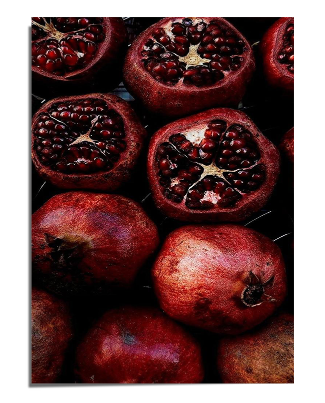 A dark and moody composition featuring multiple pomegranates, some whole and others cut open to reveal glistening red seeds. The contrast between the deep shadows and the vibrant fruit creates a dramatic and elegant visual effect.