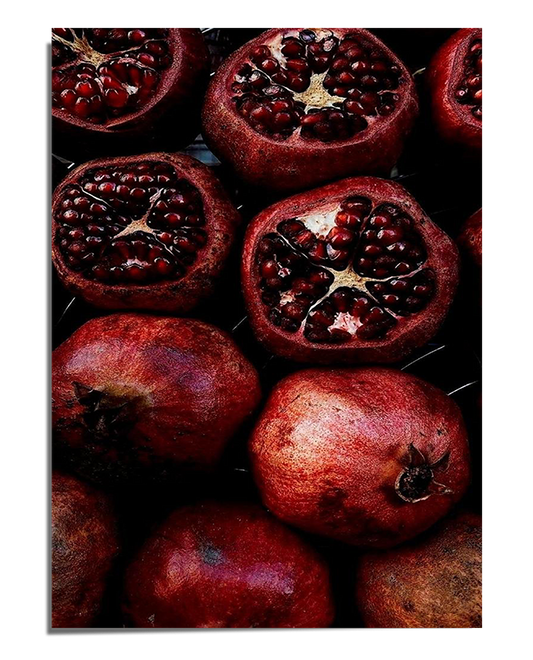 A dark and moody composition featuring multiple pomegranates, some whole and others cut open to reveal glistening red seeds. The contrast between the deep shadows and the vibrant fruit creates a dramatic and elegant visual effect.