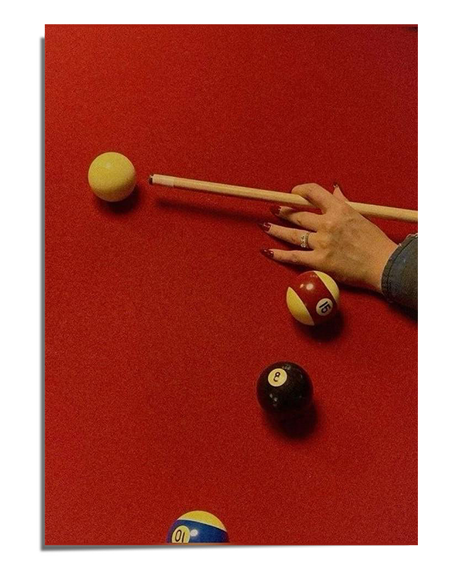 A red pool table with scattered billiard balls and a player's hand carefully positioning a shot with a cue stick. The lighting creates a warm and moody atmosphere, highlighting the texture of the felt and the contrast of the billiard balls.